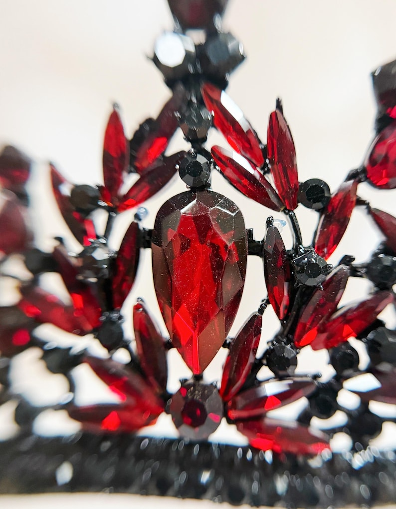 May include: Close-up of a black and red jeweled crown. The crown features a large, teardrop-shaped red gem at the center, surrounded by smaller red and black gems. The metalwork is black and intricate, creating a regal and elegant design.