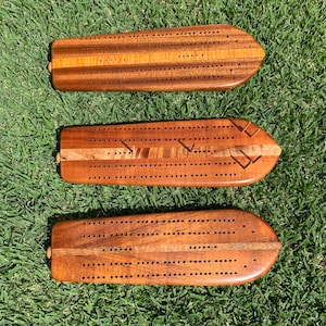 May include: Three wooden cribbage boards with a smooth, polished finish are arranged on a bed of green grass. Each board is a rich brown color with a lighter wood accent and numerous small holes for pegs. The boards have a rounded end and a small peg holder.