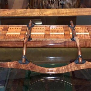 May include: A handcrafted wooden cribbage board in the shape of a canoe. The board is made of a rich, warm-toned wood with a glossy finish. It features a detailed design with pegs and paddles, and is displayed on a glass surface.