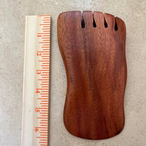 May include: A wooden jewelry display shaped like a foot, with slots for rings or other small items. The display is a rich, reddish-brown color and is approximately 6 inches long. A wooden ruler is placed next to the display.