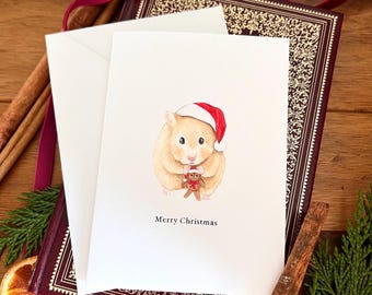 May include: A Christmas card featuring a watercolor illustration of a hamster wearing a Santa hat and holding a gingerbread man. The card is white with the words "Merry Christmas" printed below the illustration. The card is placed on a decorative book.