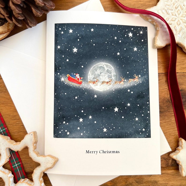 May include: A Christmas card with a watercolor illustration of Santa Claus flying in his sleigh pulled by reindeer in front of a full moon and a starry night sky. The text "Merry Christmas" is printed at the bottom of the card.