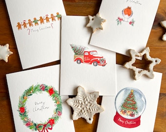 May include: A collection of Christmas greeting cards with watercolor illustrations. Designs include gingerbread men, a red truck with a Christmas tree, a robin, a wreath, and a snow globe. The words "Merry Christmas" are written on several cards.