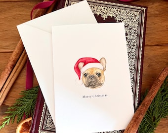 May include: A Christmas card featuring a watercolor illustration of a French Bulldog wearing a Santa hat. The card is white with the words "Merry Christmas" below the dog's portrait. The card is placed on a burgundy book with a white envelope.