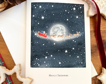 May include: A Christmas card featuring Santa Claus in a red sleigh pulled by reindeer, flying across a starry night sky with a full moon. The card has the words "Merry Christmas" at the bottom. The card is on a white background.