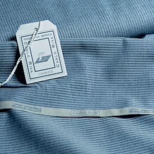 May include: Close-up of blue fabric with thin, vertical stripes. A white tag is attached with the text "CARLO RIVA" and "GALBIATE". The fabric is folded, and a strip of fabric with the text "CARLO RIVA" is visible.