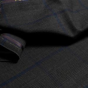 May include: Close-up of a dark gray fabric with a subtle plaid pattern and blue accents. The text "FOUR PLY TROPICAL" is visible on a dark brown band. The fabric appears to be a high-quality material, possibly for suiting.