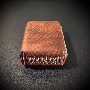 May include: A brown leather lighter case with a stitched edge. The case has a textured surface and a light brown stitching detail.