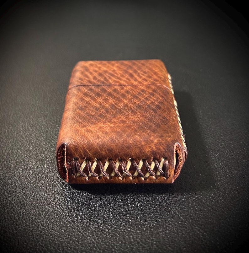 May include: A brown leather lighter case with a stitched edge. The case has a textured surface and a light brown stitching detail.
