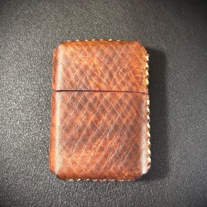 May include: A brown leather lighter case with a stitched border. The case has a textured surface and a flap that closes with a snap.