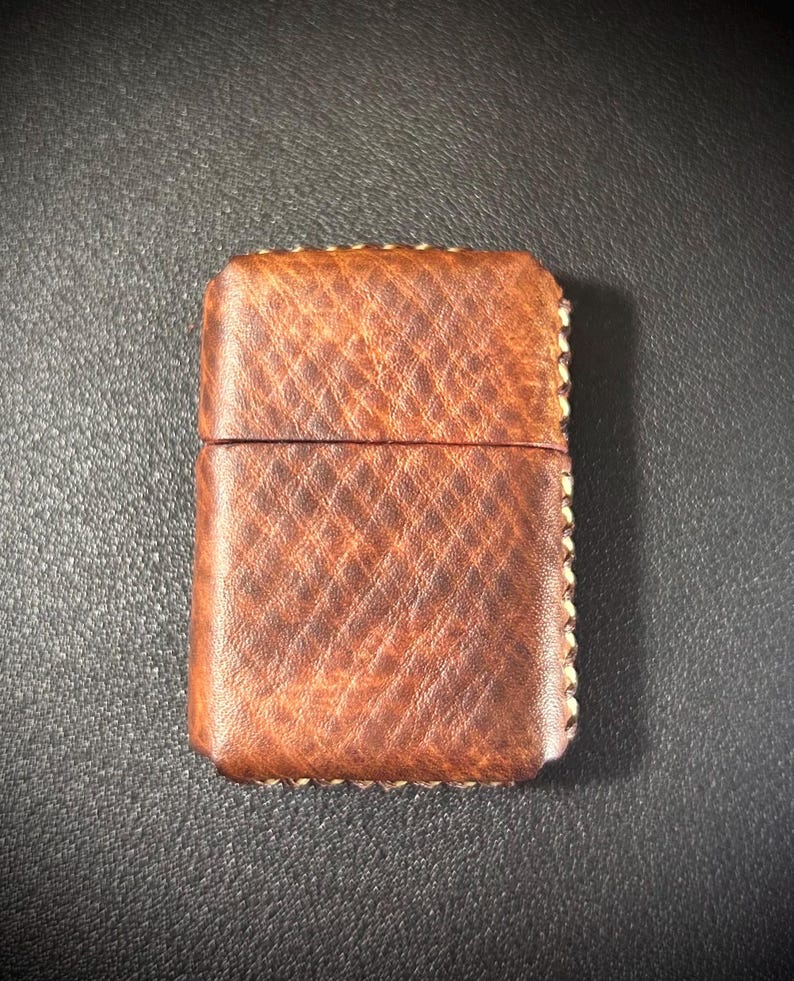 May include: A brown leather lighter case with a stitched border. The case has a textured surface and a flap that closes with a snap.