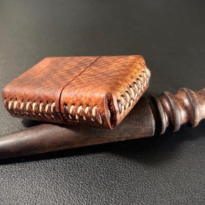 May include: A brown leather lighter case with white stitching, resting on a wooden tool with a rounded handle.