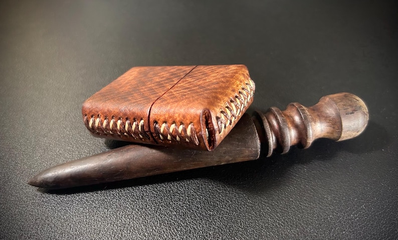 May include: A brown leather lighter case with white stitching, resting on a wooden tool with a rounded handle.
