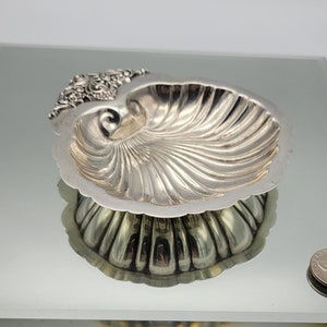 May include: A silver-colored, nautilus-shaped dish with a detailed, ridged interior. The dish has an ornate, floral-like design on one end and is reflected in the mirrored surface below. The dish is likely used for decorative purposes.