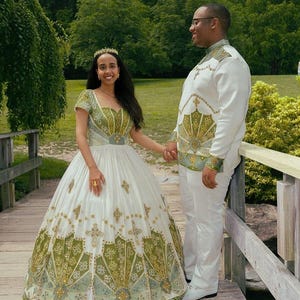 Puede incluir: Una pareja en un puente de madera, ambos vestidos con ropa formal. La mujer lleva un vestido de baile blanco con bordados en verde oliva y dorado. El hombre lleva un traje blanco con detalles a juego en verde oliva y dorado. Fondo de vegetación verde.