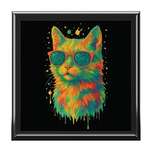 May include: A square black frame containing a colorful illustration of a cat wearing orange sunglasses. The cat is rendered in vibrant shades of yellow, orange, green, and blue with a paint drip effect against a solid black background.