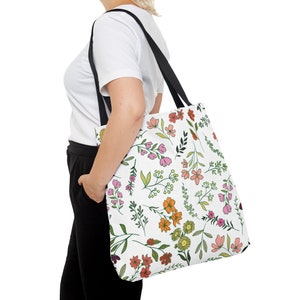 May include: White tote bag with a colorful floral pattern. The design features pink, orange, and yellow flowers with green leaves and black straps. The bag is carried over the shoulder.