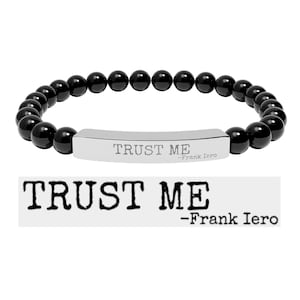 May include: A stretch bracelet featuring smooth, round, glossy black beads and a central, curved, polished silver-tone metal bar. The bar is engraved with the text "TRUST ME" in a bold, typewriter-style font, followed by "-Frank Iero" in smaller text.