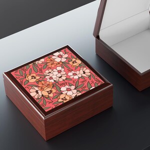 May include: A square wooden keepsake box with a dark brown finish and a hinged lid. The top of the lid features a vibrant floral pattern with pink, orange, and cream-colored flowers set against a coral background with green leaves and stems.