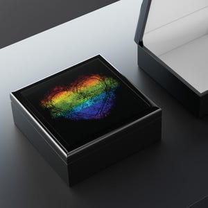 May include: A black square wooden box with a rainbow heart design on the lid. The box is closed and sitting on a gray surface.