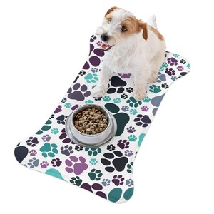 May include: A white dog food mat with a bone shape and a paw print pattern in shades of purple, teal, and gray. A small white dog is sitting on the mat, and a bowl of dog food is in front of the dog.