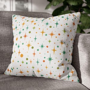 May include: A square throw pillow with a white background and a repeating pattern of colorful starbursts and dots in orange, green, and yellow. The pillow is resting on a gray sofa.