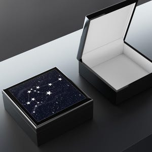 May include: A black, rectangular jewelry box with a glossy finish. The lid features a dark blue background with a white constellation design. The box is open, revealing a white interior.