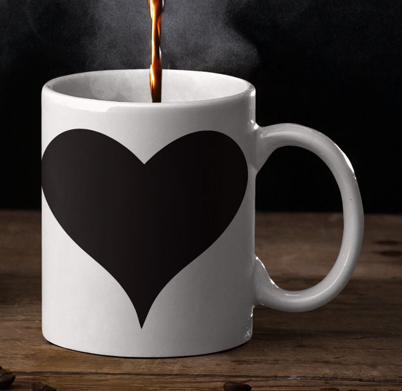 May include: A white coffee mug with a black heart design. The mug is filled with coffee and steam is rising from the mug.