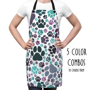 May include: An apron with a white background and a pattern of teal, purple, and black paw prints. The apron has a black neck strap and the text "5 COLOR COMBOS TO CHOOSE FROM".