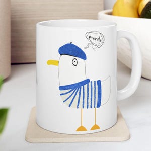 May include: A white ceramic mug featuring a graphic illustration of a white bird wearing a blue beret and a blue and white striped shirt. The bird has a yellow beak and feet, and a speech bubble above its head containing the text merde.