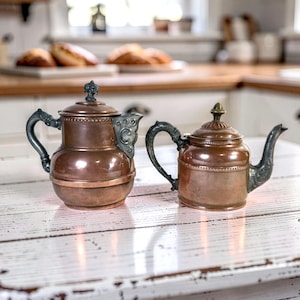 May include: Two copper teapots with metallic handles and spouts. The left teapot features a tall cylindrical body with a decorative lid finial. The right teapot has a shorter, rounded body with a pointed lid finial and ornate handle detailing.