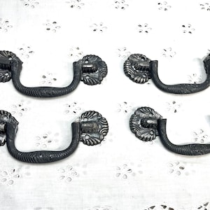 May include: Four black metal drawer pulls with ornate details. Each pull has a curved handle and a decorative, circular base.