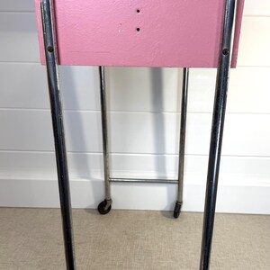 May include: A vintage pink painted cart with a rectangular top and chrome legs. The cart has small wheels for easy movement. The pink surface has two small holes. The cart is a functional and stylish piece of furniture.