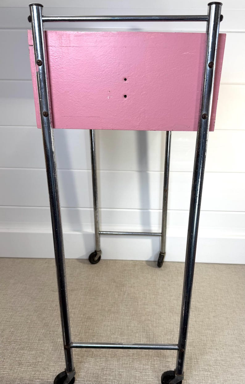 May include: A vintage pink painted cart with a rectangular top and chrome legs. The cart has small wheels for easy movement. The pink surface has two small holes. The cart is a functional and stylish piece of furniture.