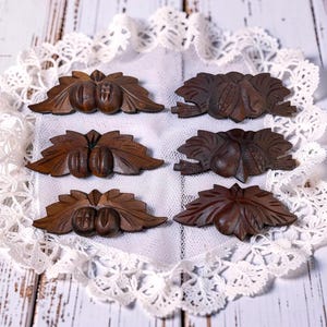 May include: Six small, intricately carved wooden ornaments. Each ornament features a pair of nuts or fruits with detailed leaf accents. The wood has a rich, dark brown color, and the ornaments are arranged on a white lace doily.