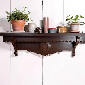 May include: A dark brown wooden wall shelf featuring a scalloped edge and a central circular carved medallion. The shelf holds two terracotta pots with green plants, a grey ceramic vase, a pinecone, a tall cylinder, and two small jars.