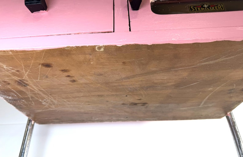 May include: A vintage pink cabinet with a wooden bottom and metal legs. The cabinet has two drawers with black handles. The word "STERILIZER" is visible on the top.