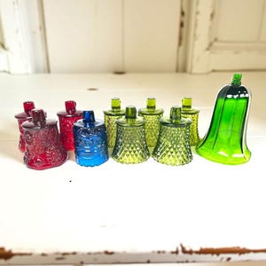 May include: A collection of colorful glass candle holders in various shades of red, blue, and green. The candle holders have a decorative design and are arranged on a white surface. One green candle holder has a bell shape.