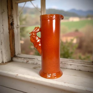May include: A tall, orange ceramic pitcher with a rooster-shaped handle. The pitcher has a glossy finish and a decorative zigzag pattern at the base. It is set against a blurred background of a window and a rural landscape.