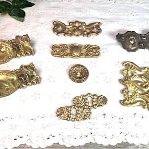 May include: Assortment of ornate brass drawer pulls and knobs with intricate designs. The pulls are oval and rectangular shaped with decorative edges. The knobs are round with a raised center.