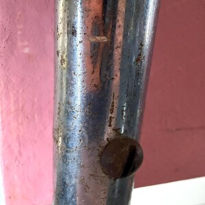 May include: Close-up of a weathered, chrome-plated metal leg with a rusty screw head. The metal has visible wear and tear, with a pink and red background.