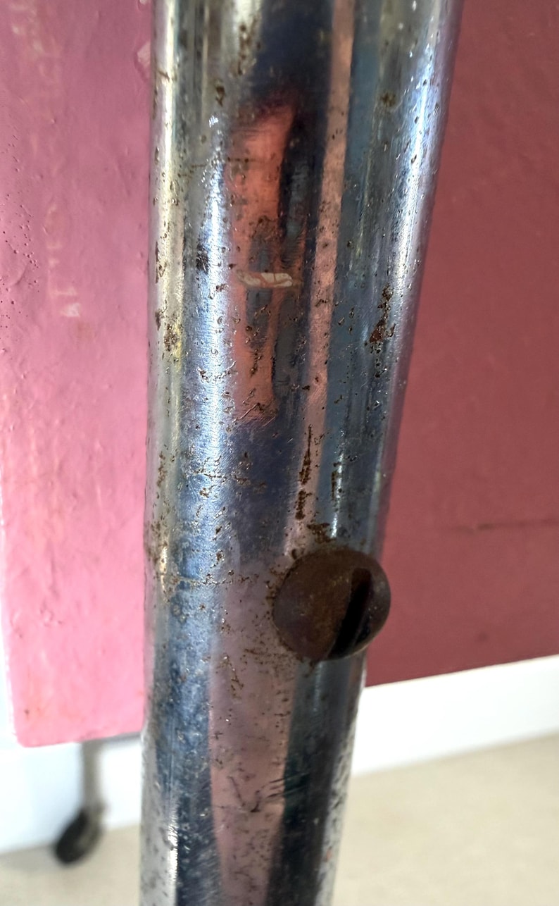 May include: Close-up of a weathered, chrome-plated metal leg with a rusty screw head. The metal has visible wear and tear, with a pink and red background.
