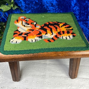 May include: A wooden footstool with a green fabric top featuring an embroidered orange and black tiger. The footstool has four legs.