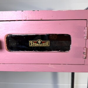 May include: A vintage pink cabinet with a black handle and a door with the word "STERILIZER" in gold lettering. The cabinet is part of a metal frame.