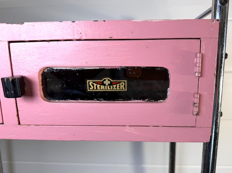 May include: A vintage pink cabinet with a black handle and a door with the word "STERILIZER" in gold lettering. The cabinet is part of a metal frame.