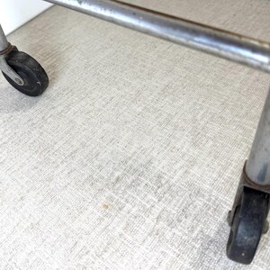 May include: Close-up of a metal cart frame with two black wheels. The frame is made of silver metal and has a rectangular shape. The wheels are black and appear to be made of rubber. The cart is on a light-colored surface.