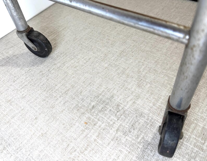 May include: Close-up of a metal cart frame with two black wheels. The frame is made of silver metal and has a rectangular shape. The wheels are black and appear to be made of rubber. The cart is on a light-colored surface.