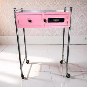 May include: A vintage pink metal cabinet with two drawers and a door, mounted on a chrome frame with wheels. The cabinet has black handles and the word "Sterilizer" is visible on the door.