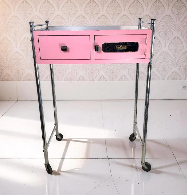 May include: A vintage pink metal cabinet with two drawers and a door, mounted on a chrome frame with wheels. The cabinet has black handles and the word "Sterilizer" is visible on the door.
