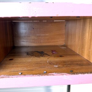 May include: An open wooden box with a pink painted exterior. The interior is natural wood with visible grain. The box appears to be part of a larger piece of furniture, with metal hardware visible on the sides.